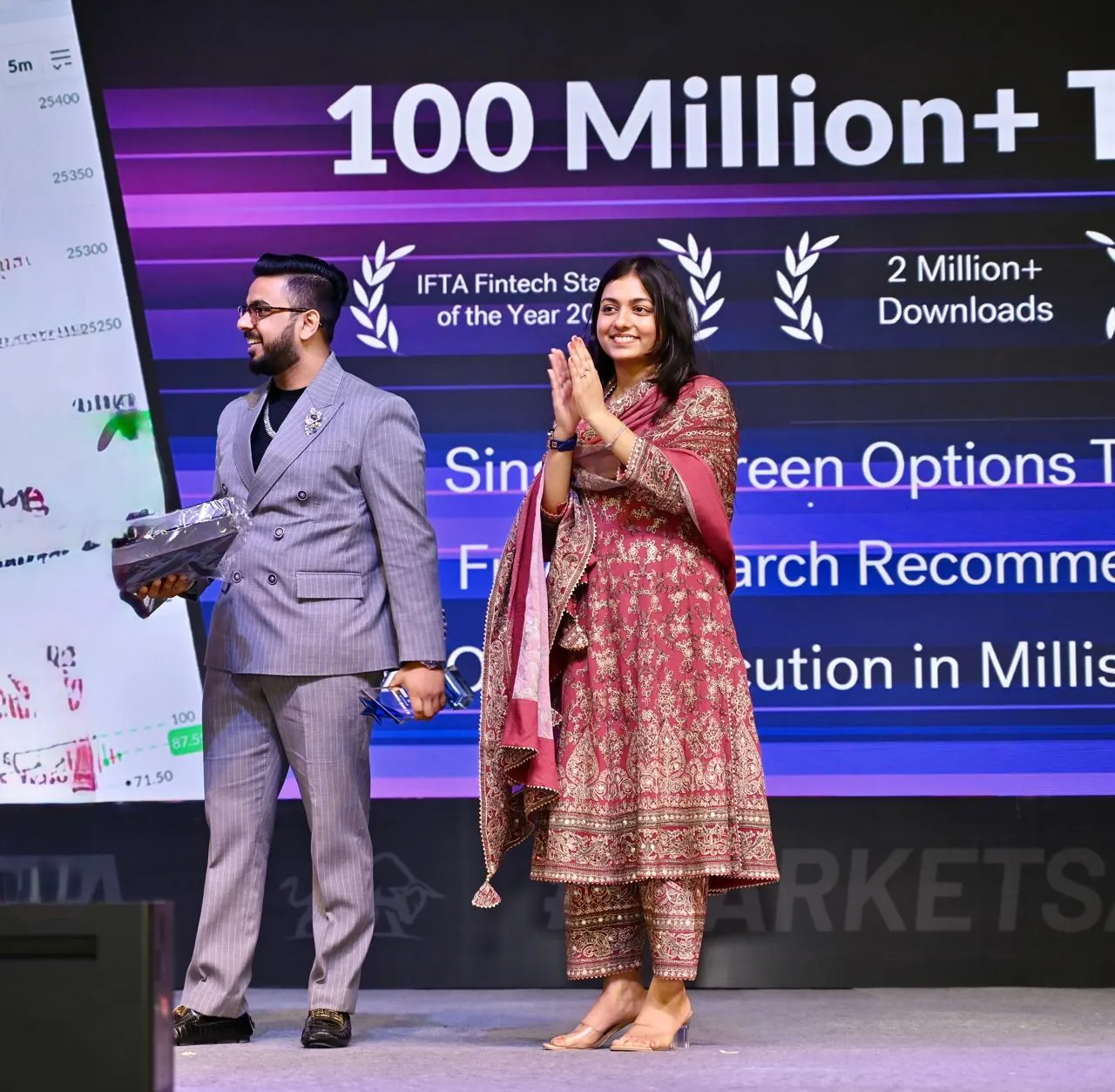 Two presenters on stage beside a screen showing platform milestones