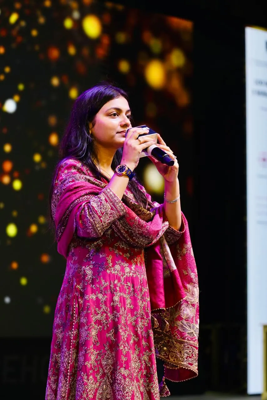 A speaker addresses the room with a handheld mic under stage lights