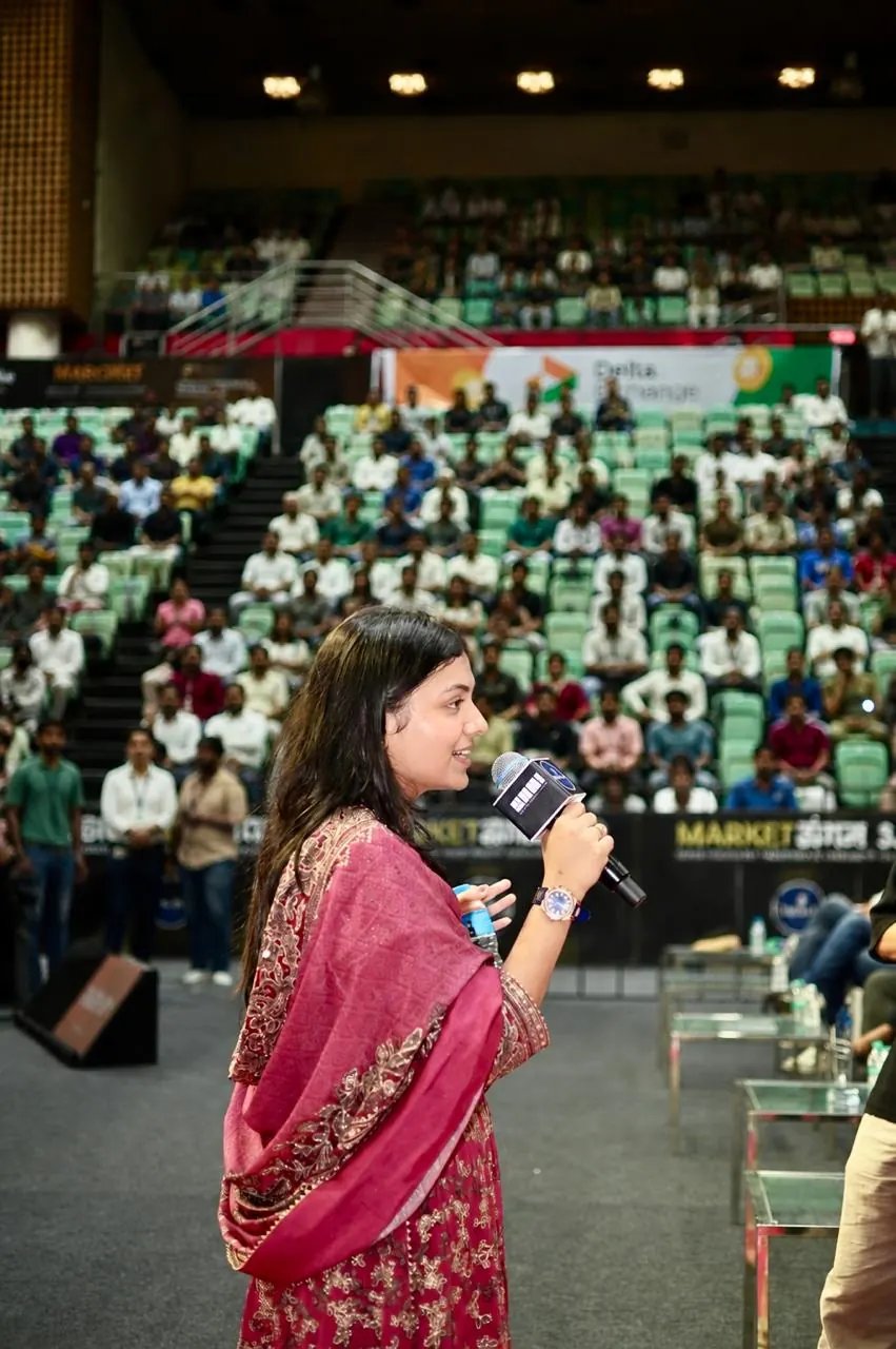 A speaker holds a mic in the aisle, a packed auditorium behind